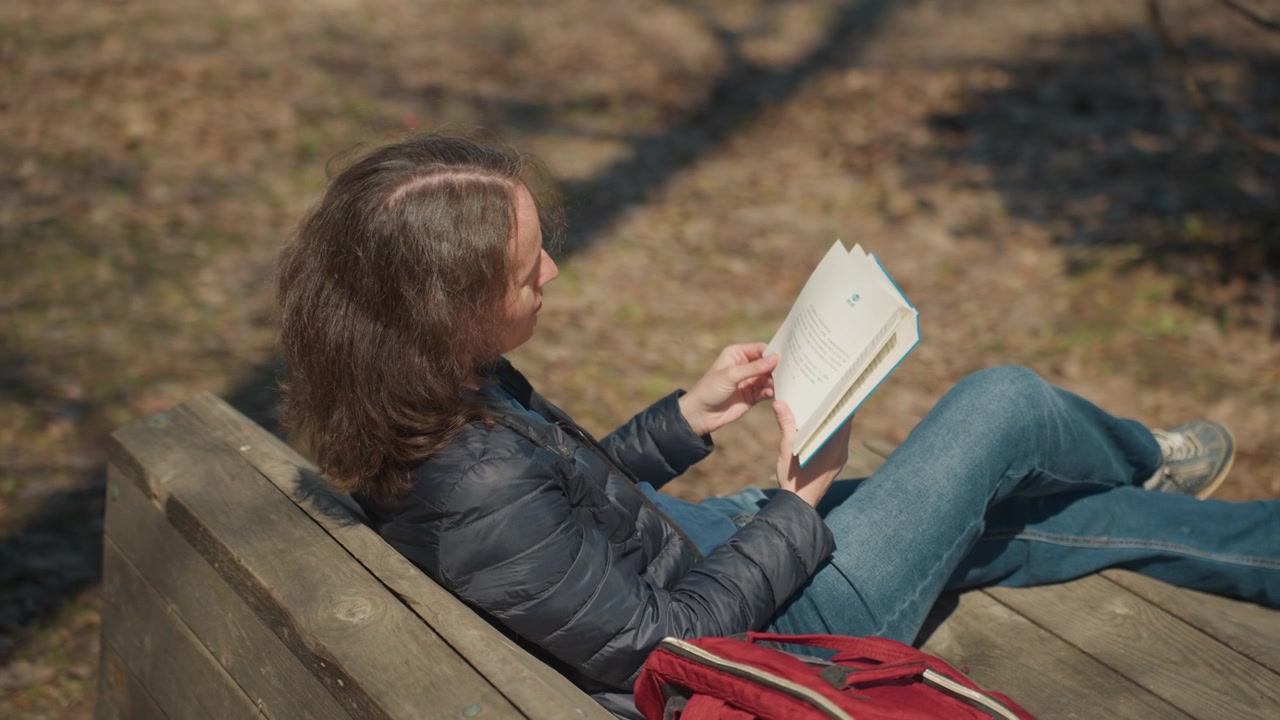 Una donna con i capelli lunghi legge un libro mentre è seduta su una panchina di legno in un parco, indossando una giacca scura e jeans. Il libro è aperto mentre la donna lo tiene, una borsa rossa è appoggiata lì vicino. La luce del sole crea ombre.