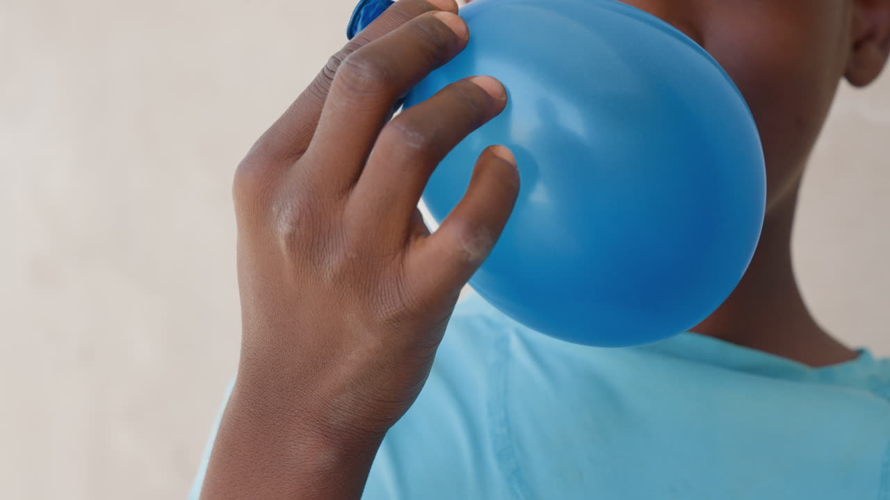 Child with blue shirt blowing balloon closeup, fingers gripping knot with sand on hands, shirt fabric and sleeve visible, subtle smile and anticipation for outdoor game or celebration, candid street