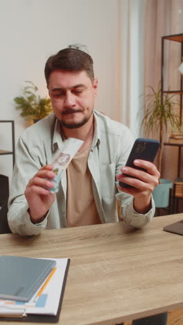 smiling caucasian young man counting money cash using smartphone at home office table in living room