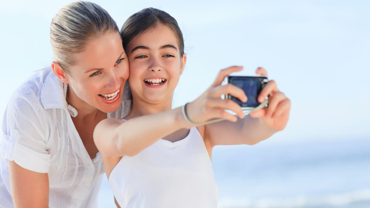 retrato de una madre y una hija caucásicas sonrientes de vacaciones tomando fotos en el mar