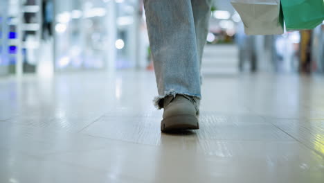 lower angle leg view of someone in expensive shoes and jeans walking in a well-lit mall with shopping bag, blurred figures moving and blurry lights in background