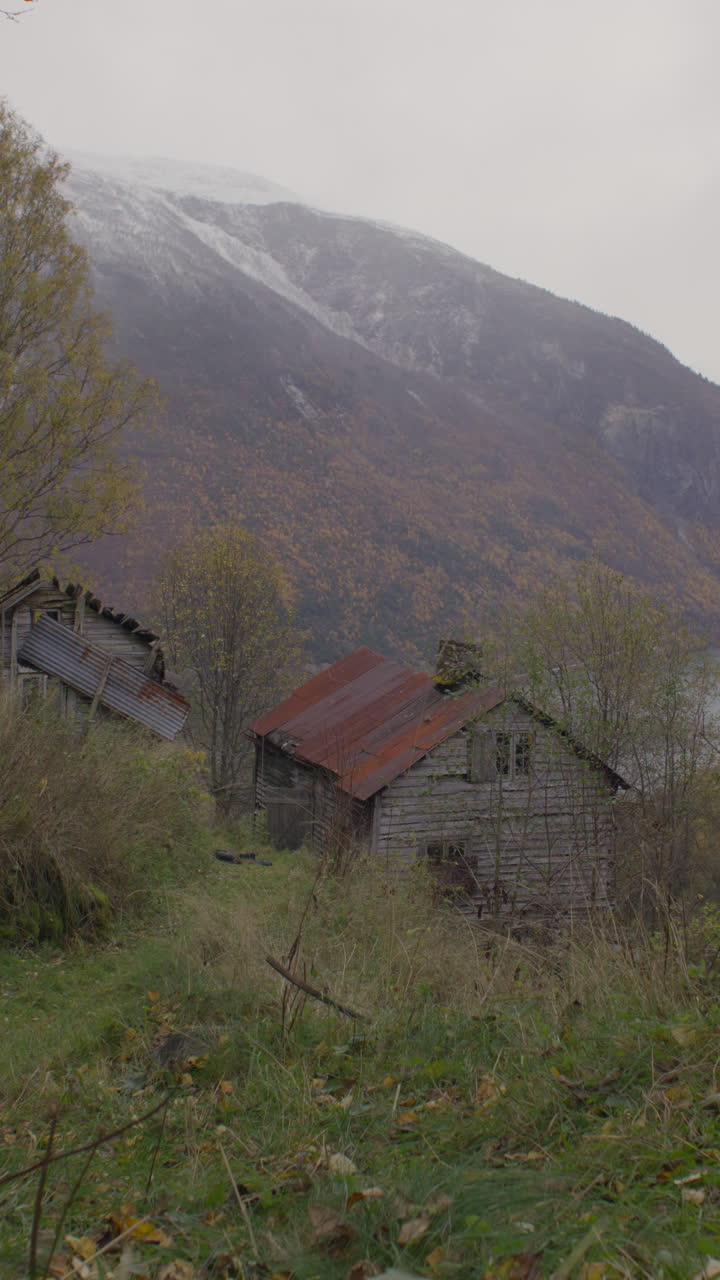 Vertical shot of old wooden houses with rusted roofs stand abandoned in a remote valley, with autumn trees and snowy mountains. Astonishing view.