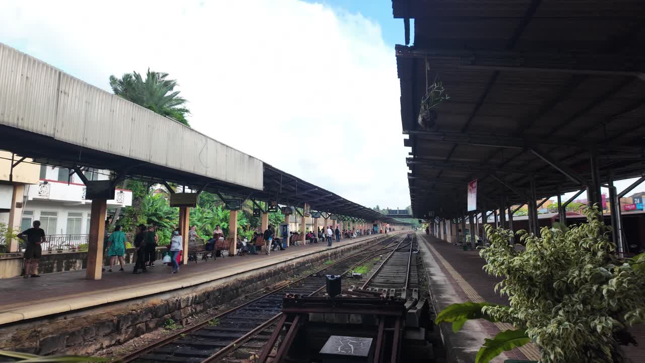 Kandy Railway Station in Sri Lanka, where people are gathered on the platform waiting for their train. The empty tracks stretch into the distance under a covered roof. Pan Right