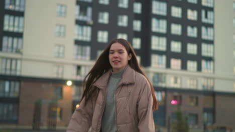 a young woman in a peach jacket and black trousers rollerblades with a joyful expression through a modern urban area, with tall residential buildings in the background. her hands are raised