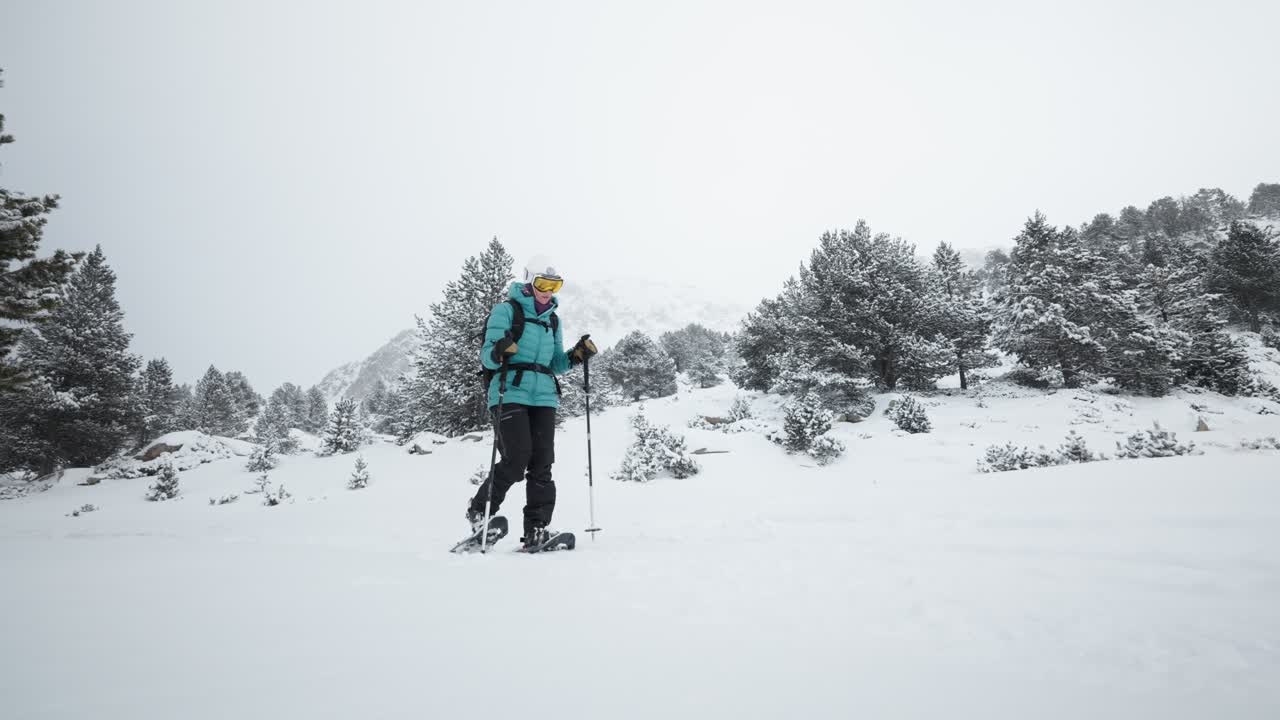 Low-angle shot of a snowshoer with yellow ski mask walking in snowfall. Mountains and snowy trees in Andorra.