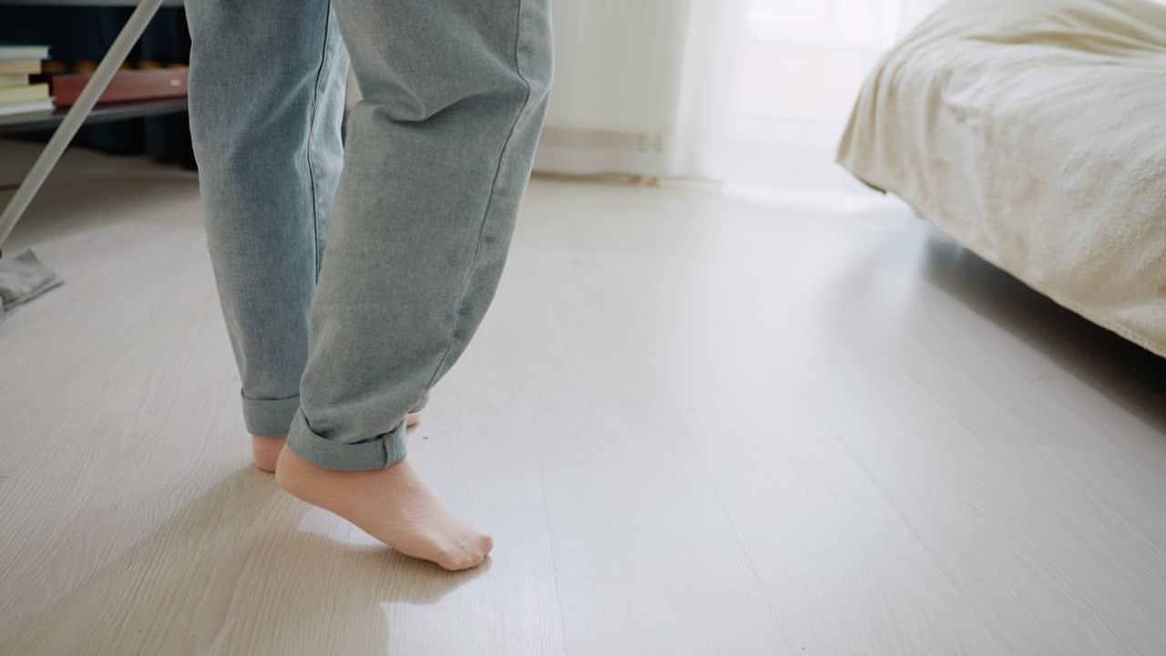 Lower angle view of cleaner using mop to wash wooden floor while another person tidies bed, showcasing daily household chores, hygiene, and cooperation in bright cozy living room environment