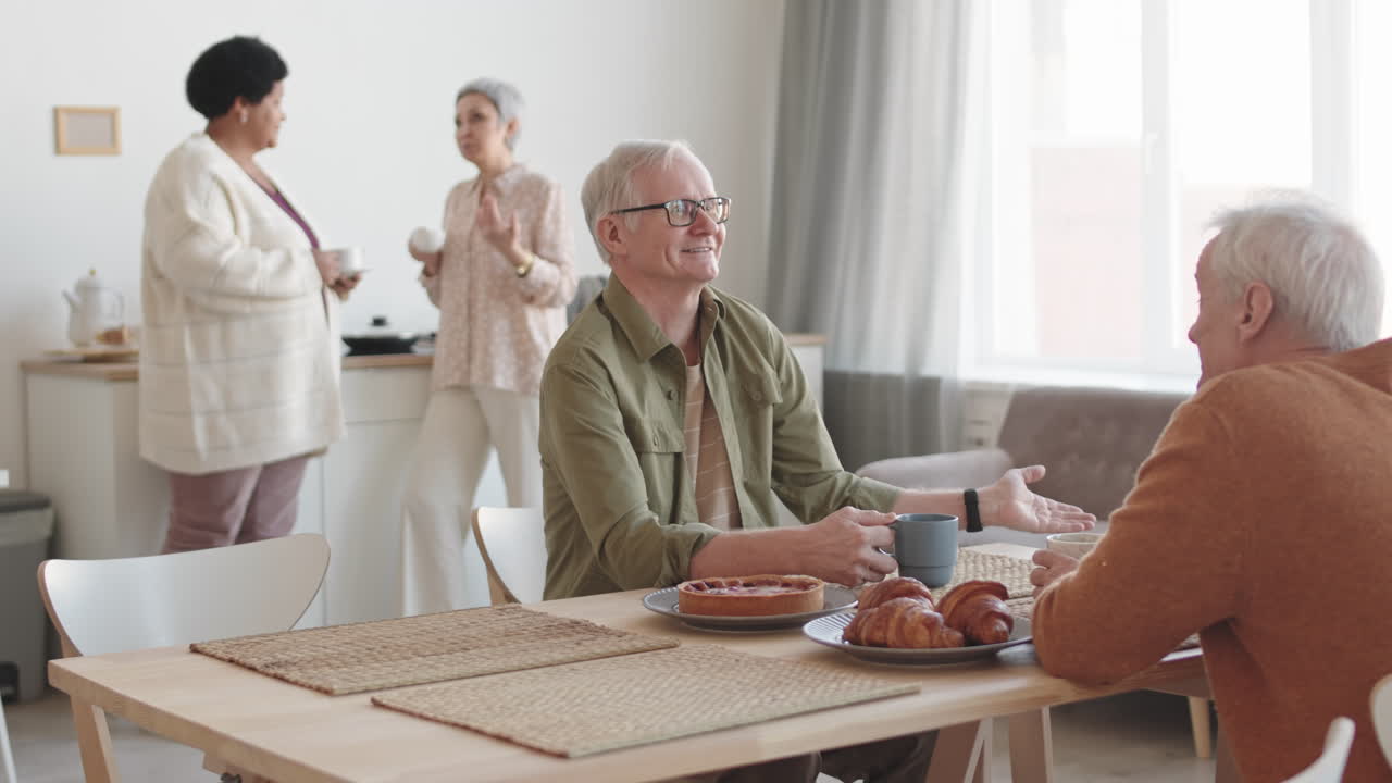 Medium long of cheerful Caucasian older man sitting at table, having conversation over tea with friend, multiethnic women joining them, coming from background