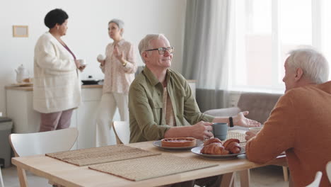 medium long of cheerful caucasian older man sitting at table, having conversation over tea with friend, multiethnic women joining them, coming from background
