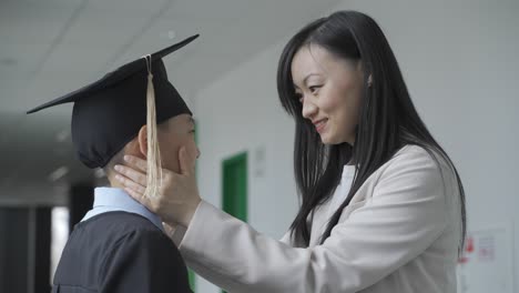 asian mother touching her son's face. he is wearing a gown and a mortarboard.