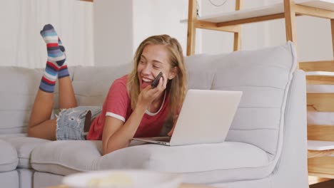 hermosa mujer hablando en un teléfono inteligente y usando una computadora portátil mientras está acostada en el sofá