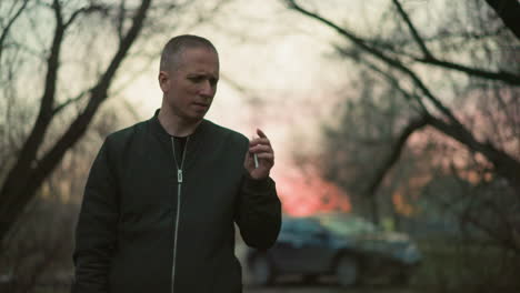 un hombre con una chaqueta verde de pie al aire libre, sosteniendo un cigarrillo y contemplando, con un fondo borroso con árboles y un coche