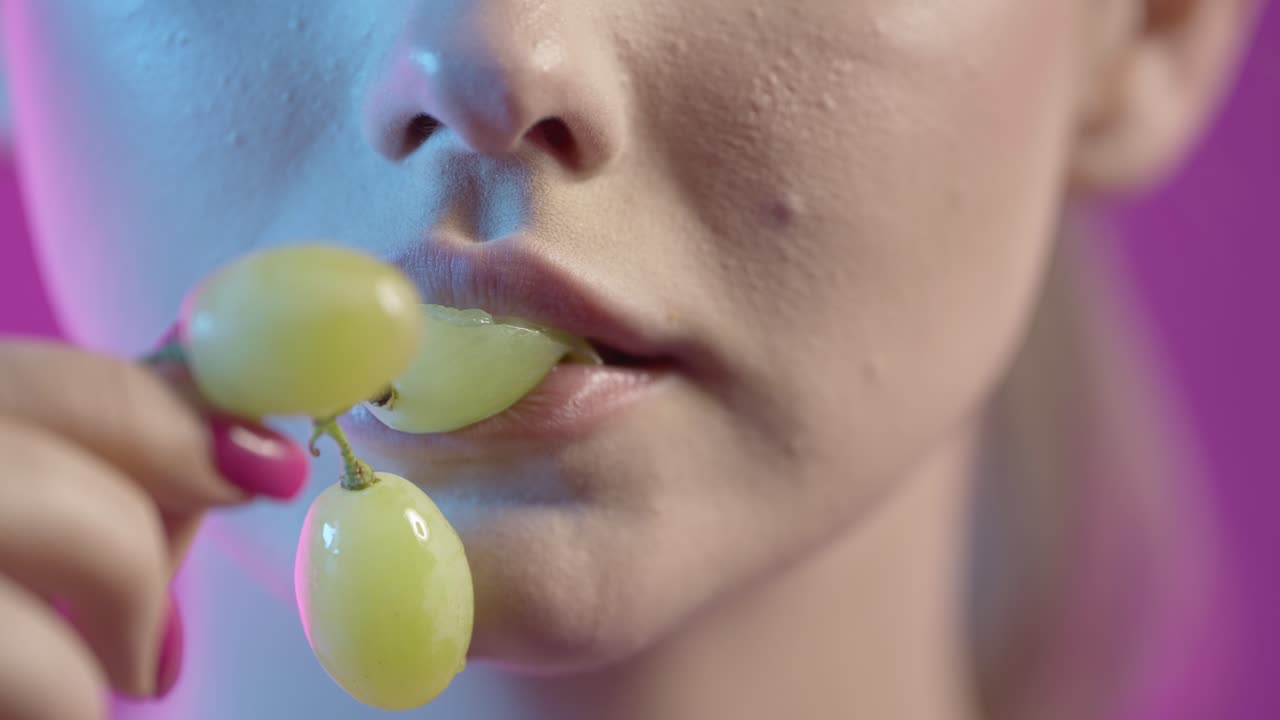 primer plano de la cara y la boca de la mujer, comiendo uvas verdes contra un fondo púrpura, toma de estudio