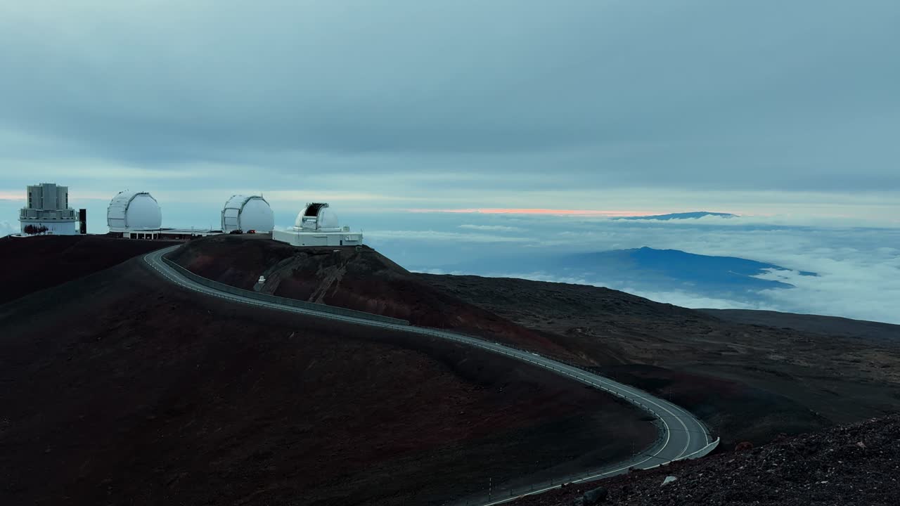 A mountain road curves toward Mauna Kea observatories above cloud-covered terrain