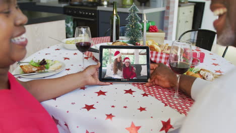 una pareja afroamericana sonriente usando una tableta para una videollamada de navidad con una familia feliz en la pantalla