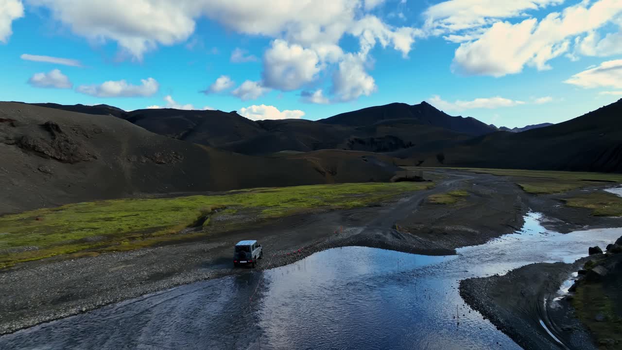 A 4×4 navigates a shallow river crossing in the Icelandic highlands, driving toward a wide valley of dark volcanic slopes under bright summer clouds
