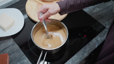 person stirring smooth golden morn porridge on electric stove, creating a rich, creamy texture, partial view of a white plate and wooden cutting board visible on a dark kitchen countertop