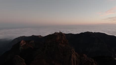 drone footage showing hikers standing atop a rugged summit surrounded by a vast cloud inversion during early sunrise in madeira.