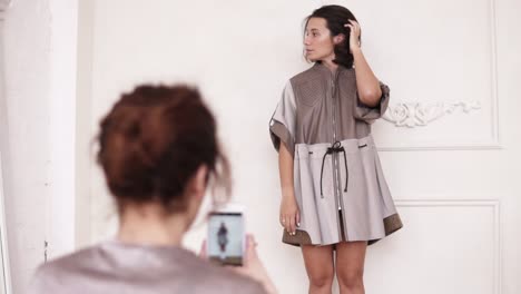 Unrecognizable-girl-with-hair-pulled-in-a-bun,-taking-picture-with-a-smartphone-of-a-young-stylish-girl,-wearing-elegant-leather-dress,-standing-on-white-wall-background