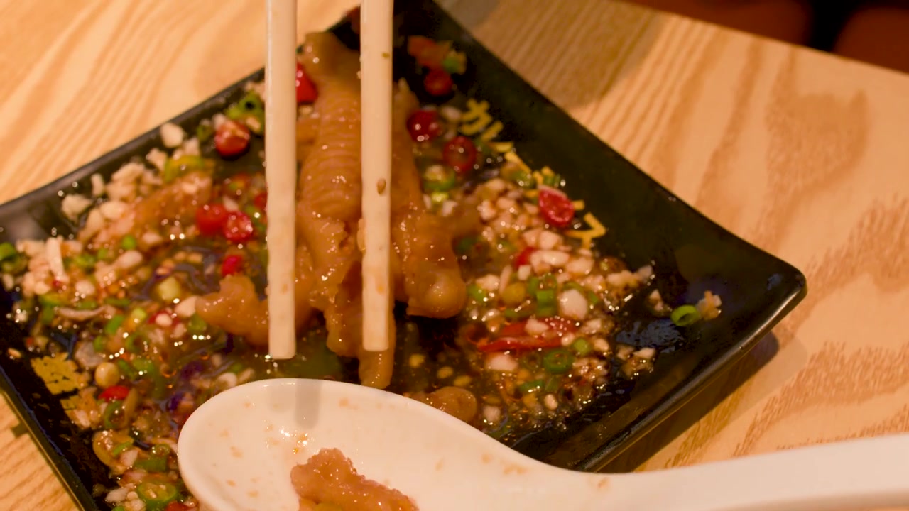 Static close-up shot of chopsticks picking up spicy chicken feet from a black ceramic plate.