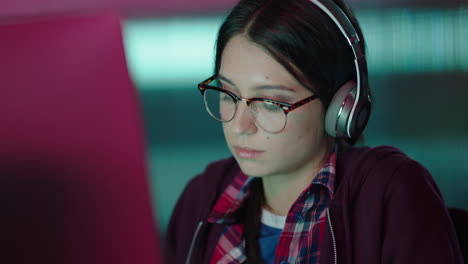 young caucasian woman student working late using computer typing research project browsing online brainstorming deadline in trendy office at night listening to music wearing headphones