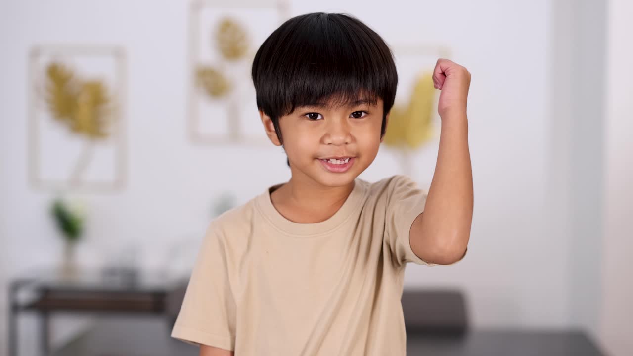 Young Asian boy in beige shirt smiles and gives a thumbs up gesture in a bright, modern room with soft, even lighting and a static camera