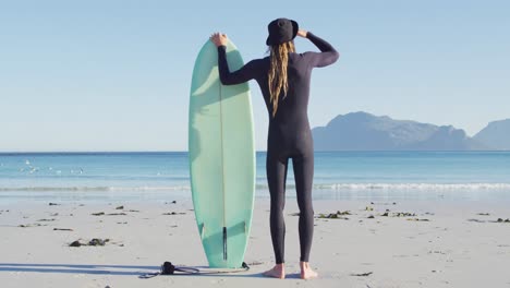 Video-Der-Rückansicht-Eines-Kaukasischen-Mannes-Mit-Dreadlocks-Im-Neoprenanzug,-Der-Ein-Surfbrett-Am-Sonnigen-Strand-Hält