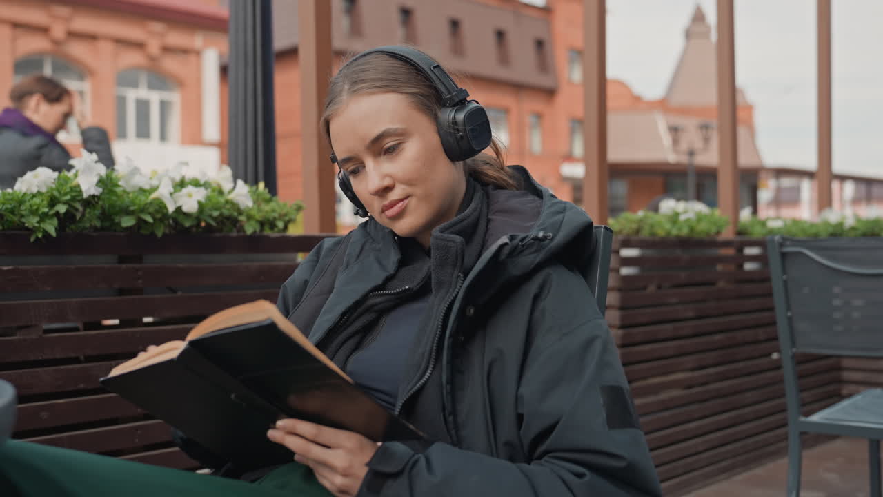 Ein Leser im Straßencafé mit Kopfhörern sitzt mit einem To-Go-Becher auf der Terrasse und blättert in einem abgenutzten Roman, eingehüllt in eine dunkle Jacke, urbane Backsteingebäude im Hintergrund, Blumenkästen in der Nähe, sanft