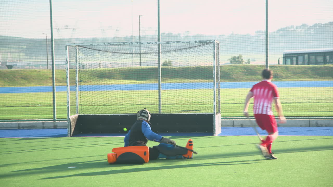 Playing field hockey, goalie in protective gear blocking shot from opponent