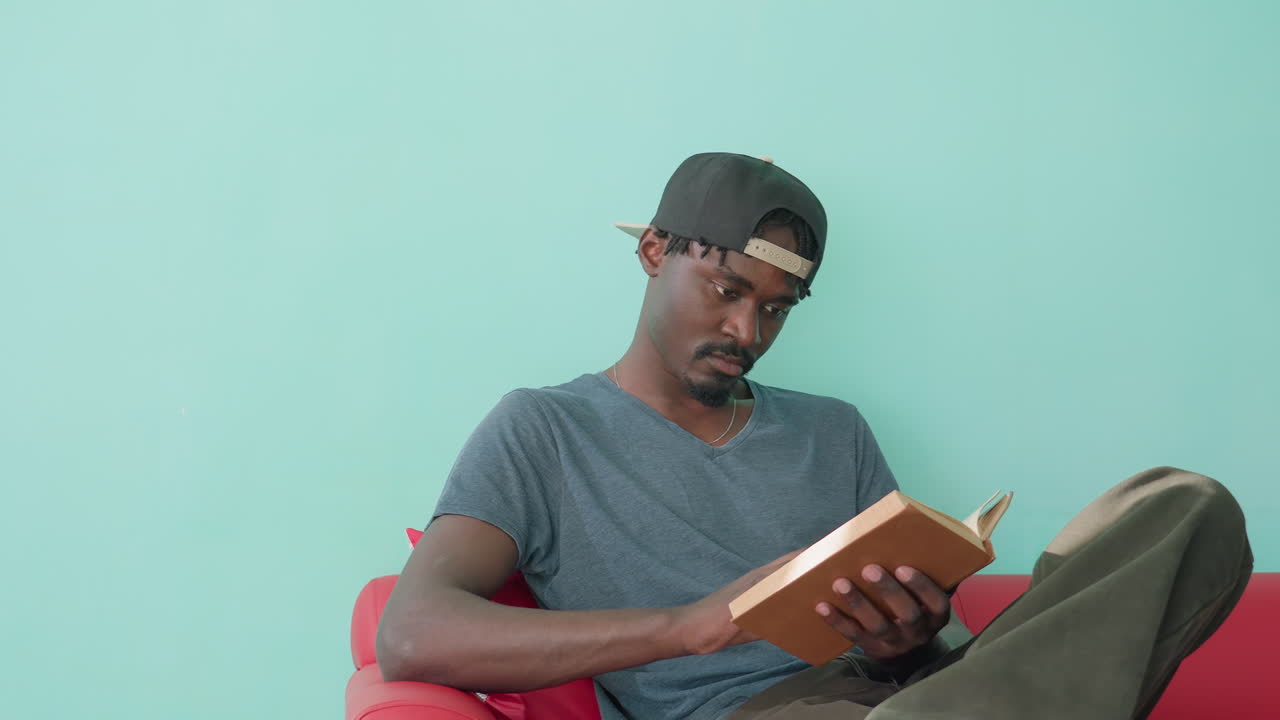 Jeune homme en tenue décontractée, assis sur un canapé rouge avec une jambe relevée, feuilletant les pages d’un livre qu’il tient entre les mains, adossé à un mur turquoise, semblant absorbé par une activité de lecture intérieure paisible et réfléchie.