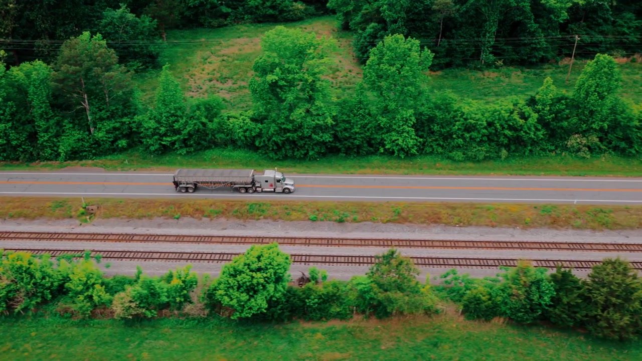 Side aerial of an open-top bulk commodity trailer under a rolling tarp, hauled by semi along a quiet rural highway parallel to railroad tracks.