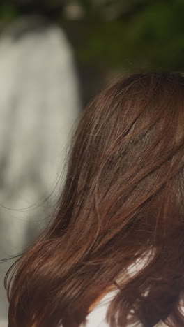 woman with messy brown hair against blurry waterfall at hike closeup. tourist observes heavy water flow falling from steep cliff on summer vacation