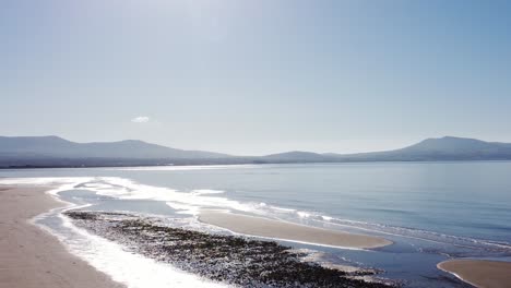 aerial view newborough beach sand dune coastline and sparkling seascape with misty snowdonia mountain range skyline, rising pan left