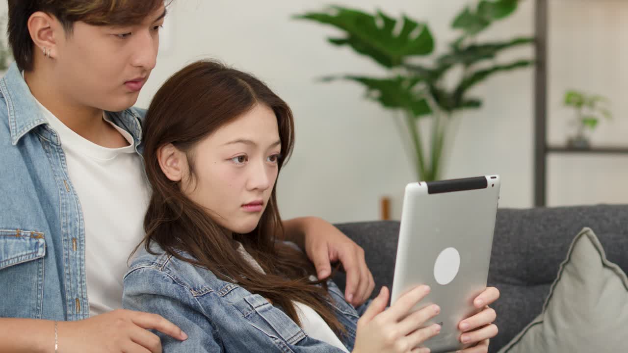 明るいリビングでタブレットを見ている若いアジア人のカップルが、突然驚いたように反応している。