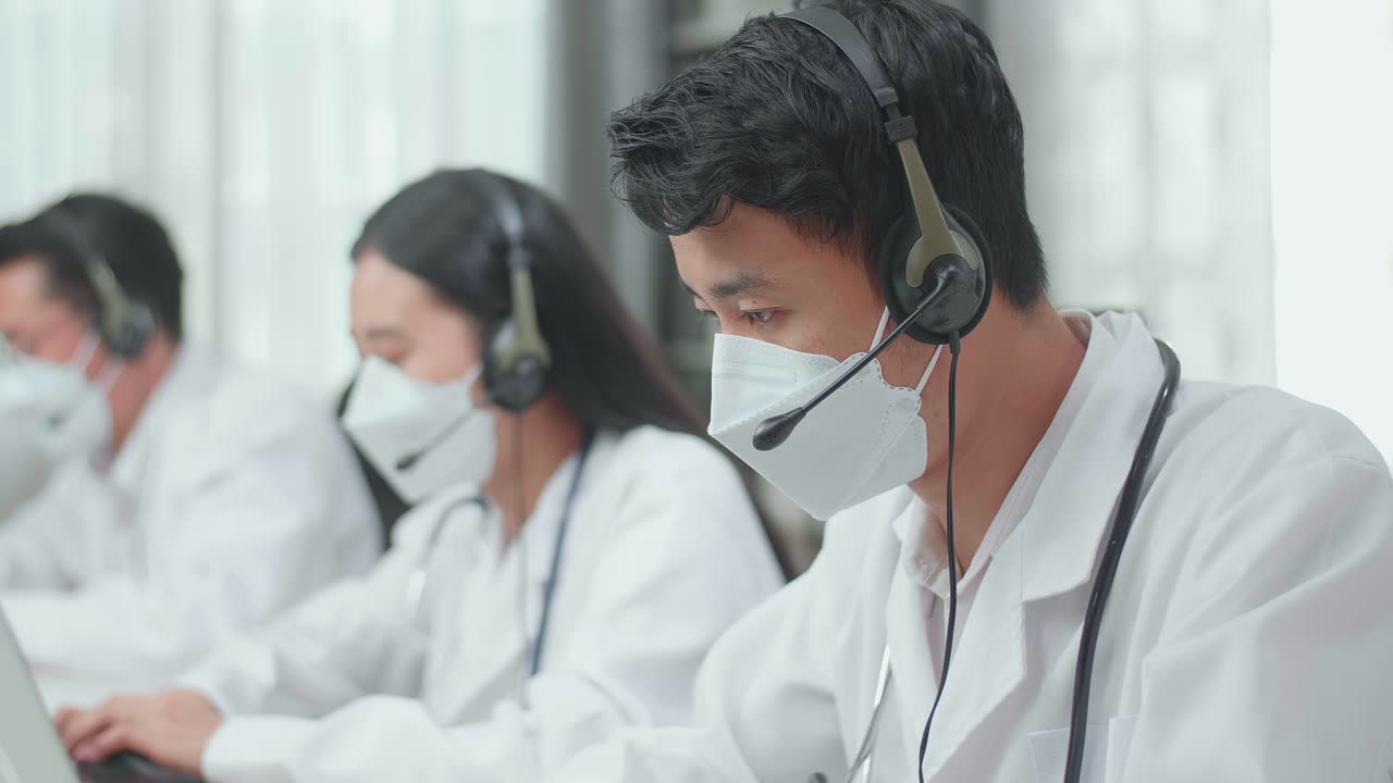Three Asian Doctors Wearing Headsets And Masks Working As Call Centre Agent Speaking To Customer On The Call And Typing During Woking With Colleagues At The Office
