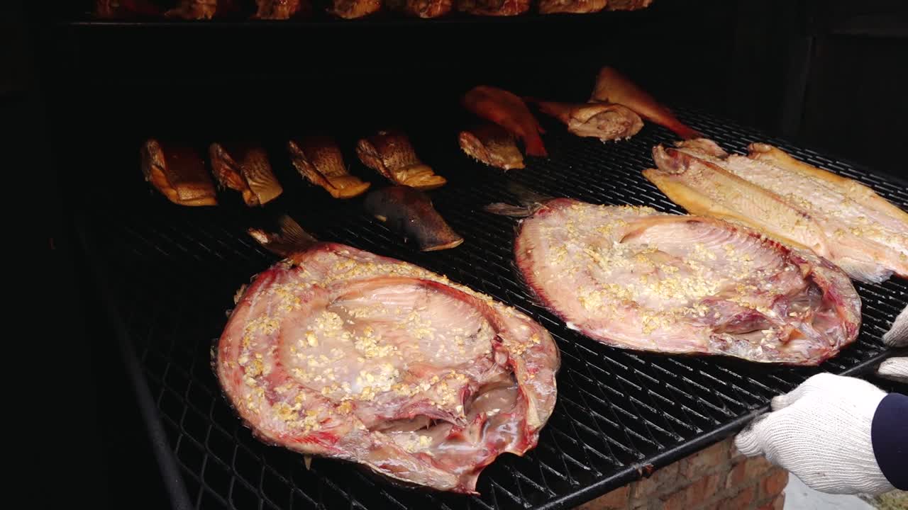 Various types of fish, including eel and redfish, are being smoked on a grill rack. The golden-brown color and rich texture highlight the traditional smoking process.