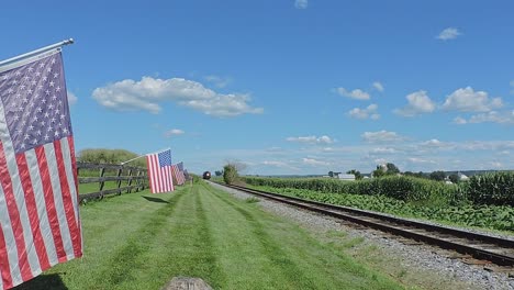 a restored antique steam passenger train approaches a fence with gently waving american flags it, on a sunny summer day