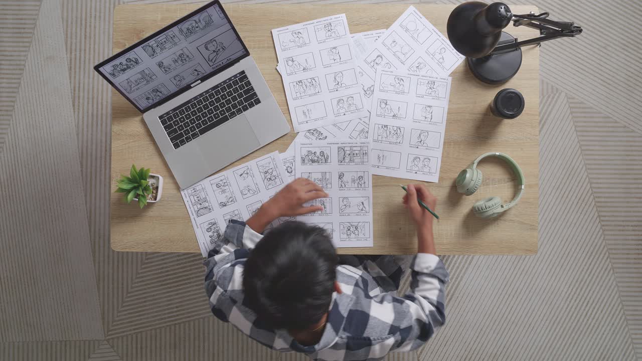 vista superior del artista masculino asiático entrando en el estudio luego sentado y dibujando el guión gráfico para la película en la mesa con una computadora portátil y auriculares