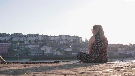 back view of a blonde girl sitting on the quay looking in the distance with st ives in the background on a sunset in cornwall, england, uk