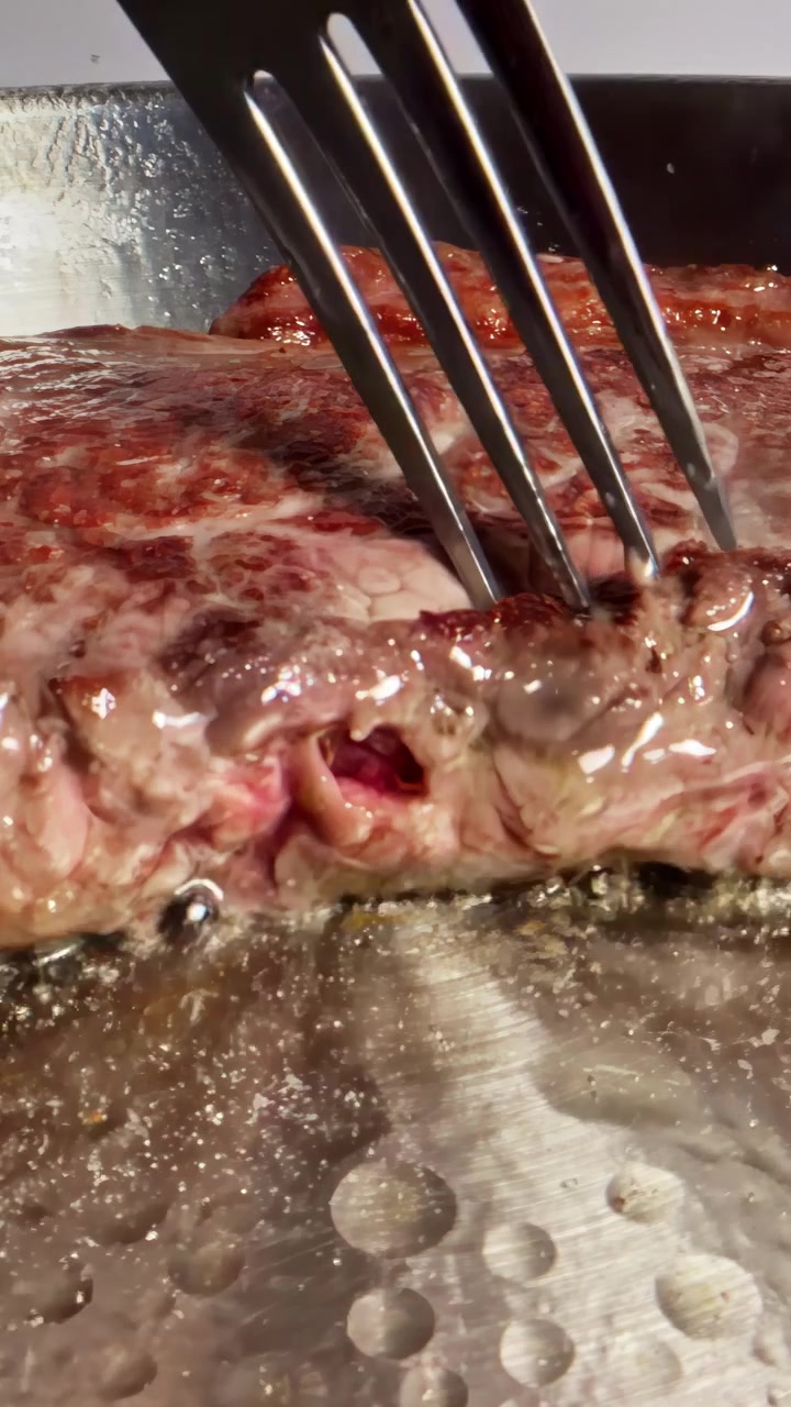 Forchetta di metallo che gira una bistecca di manzo cruda in una padella rovente con burro e succhi di carne, processo di cottura al sangue, ripresa statica in primissimo piano.