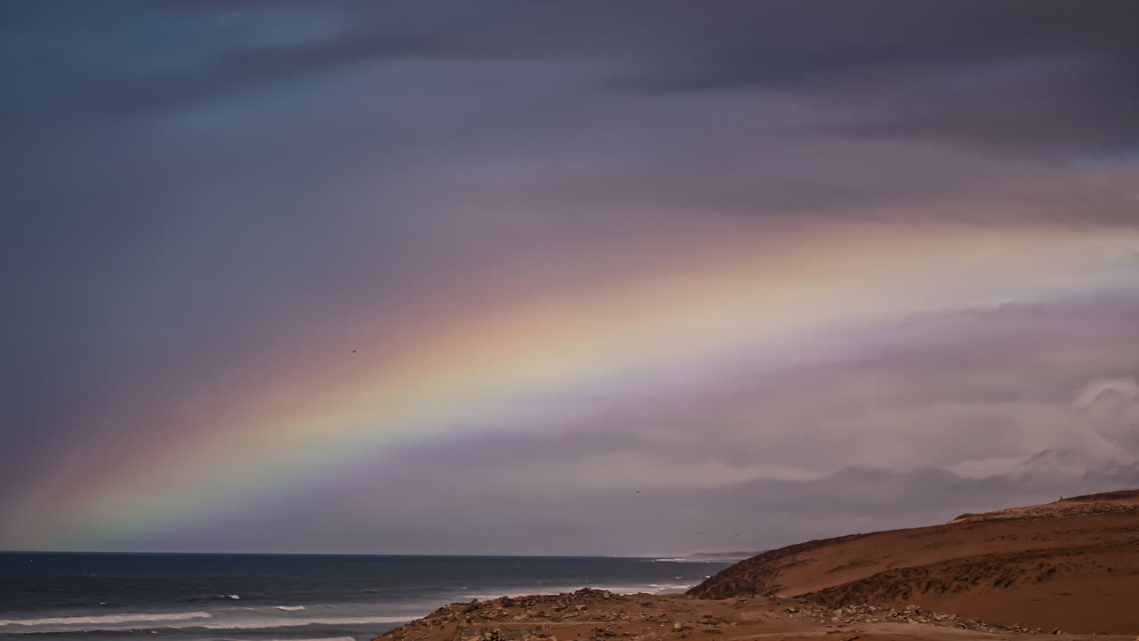 tiro de lapso de tiempo de la iluminación del arco iris de colores sobre el océano y la playa de arena durante el día nublado y soleado