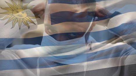 Animation-of-flag-of-uruguay-waving-over-man-wearing-face-mask-during-covid-19-pandemic