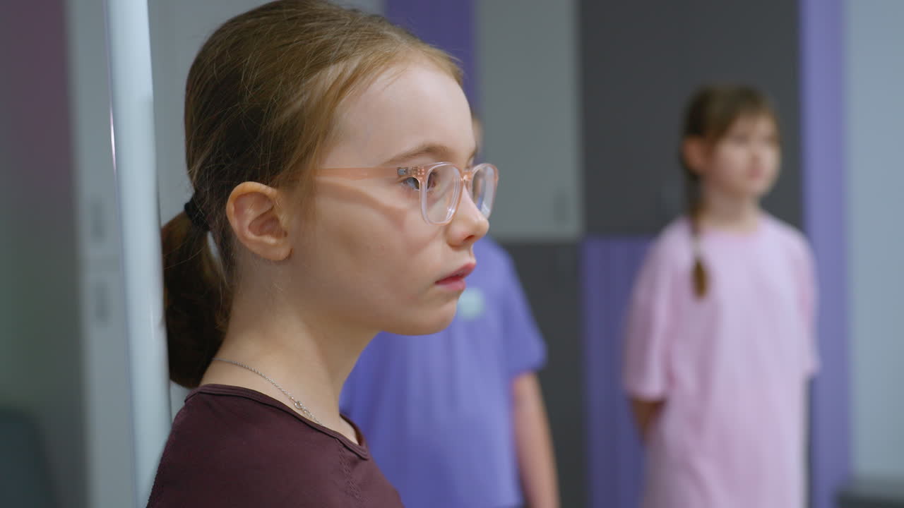 안경을 입은 어린 소녀가 기둥에 앉아있는 클로즈업, 배경에 다른 두 소녀를 관찰하는 동안, 분홍색을 입고 상호 작용하는 장면, 장면은 체육관 또는 훈련 시설에서 실내에서 설정됩니다.