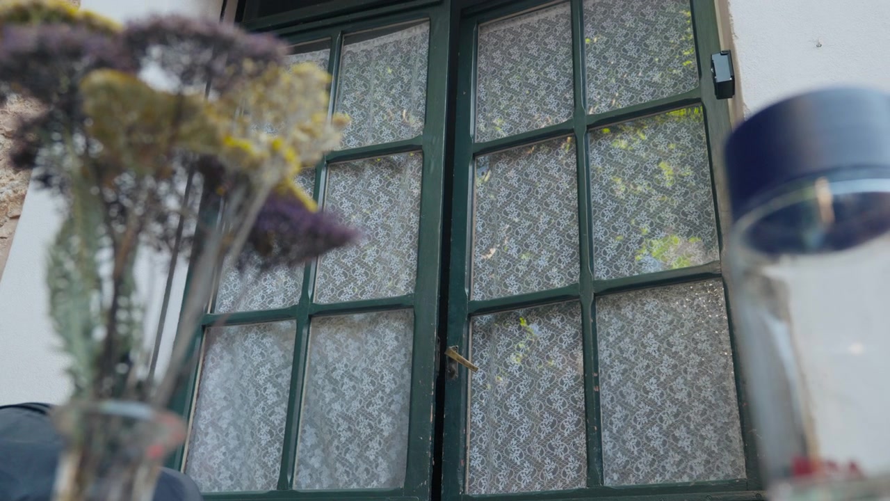 An old green multi-pane window with white lace curtains, dried wildflowers soft in the foreground. Nostalgic Mediterranean house detail for lifestyle edits. Medium shot.