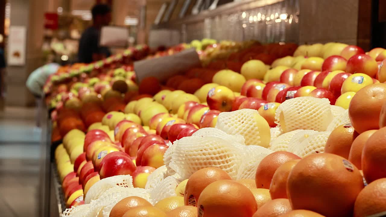 Colorful fruits arranged in a supermarket aisle with soft lighting and a passing shopper