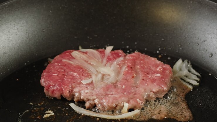 Memasak Patty Hamburger Mentah dengan Bawang di Wajan.