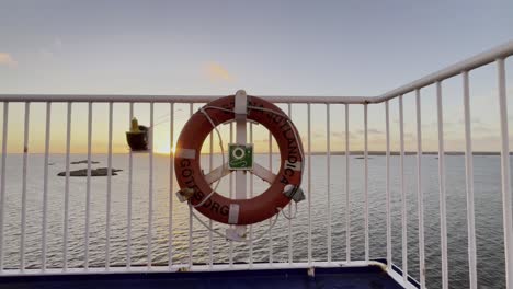 Lifebuoy-on-the-railing-of-a-car-ferry-with-some-stone-islands-in-the-background-in-the-sea-in-the-evening