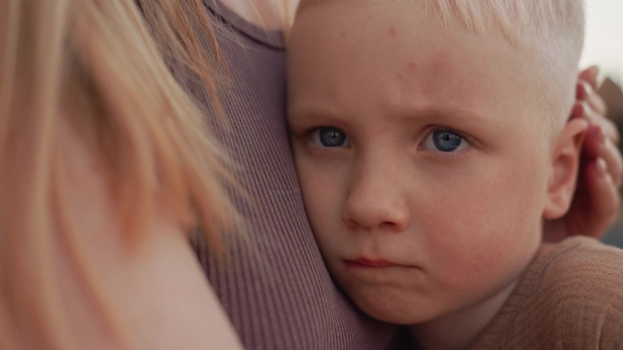 Un bambino con i capelli chiari è tenuto stretto da una donna, entrambi in una luce naturale soffusa. La scena si concentra sulla loro connessione intima e sulla texture dei dettagli dell'abbigliamento, come il tessuto a costine e la maglieria.