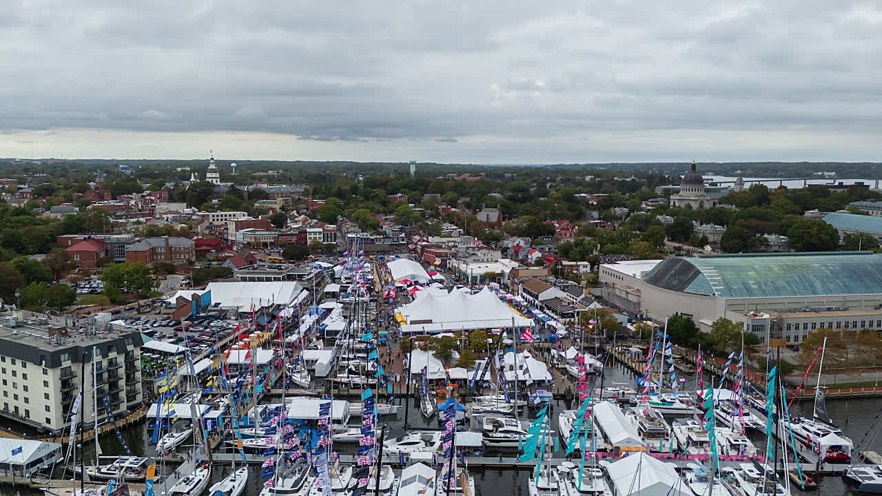 Annapolis Boat Show. En livlig festival vid vattnet fyller marinan, med rader av båtar, färgstarka flaggor och tält mot en stadsbild under en molnig himmel - Hyperlapse