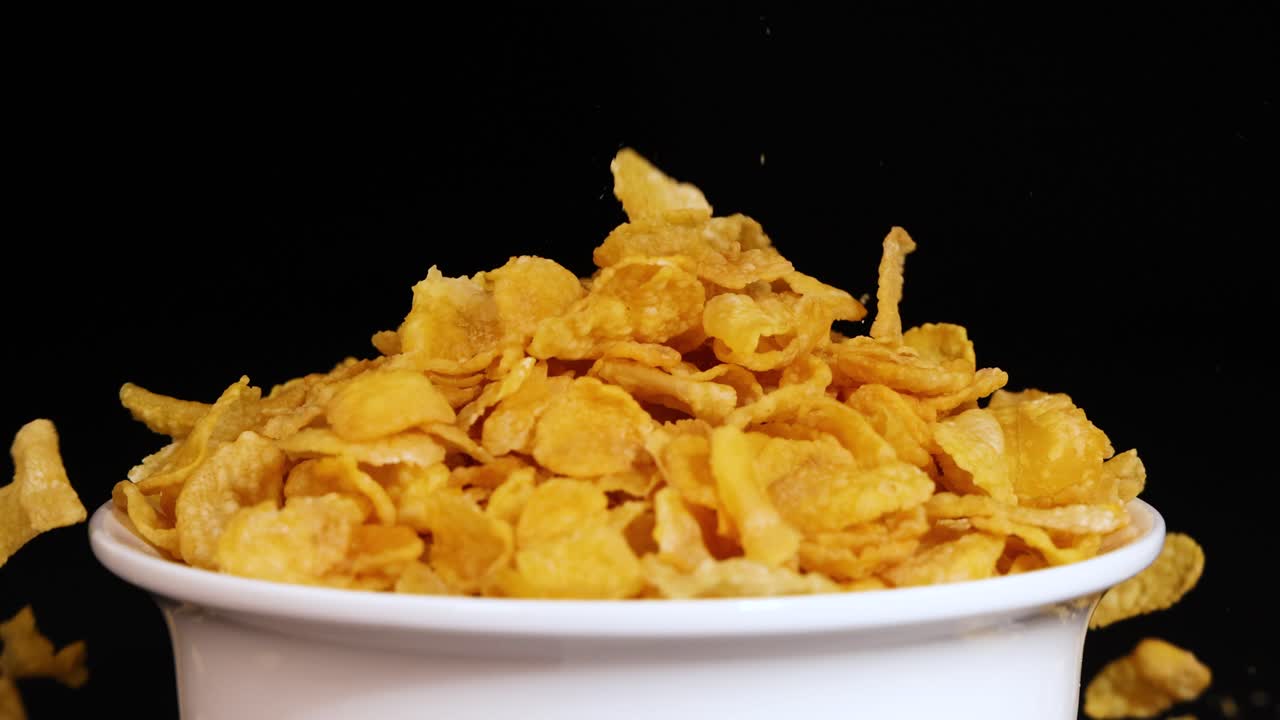Cornflakes cascade into a white bowl, set against a black backdrop, highlighting the golden cereal in motion