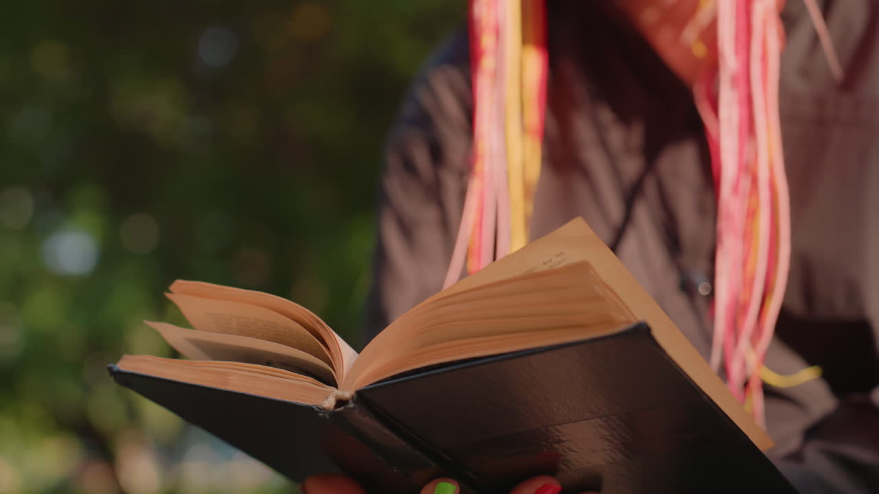 Persona leyendo una novela abierta bajo los árboles en un banco, sombra suave y reflejos cálidos, dedos sujetando el lomo y pasando las páginas, chaqueta informal y mechones de pelo coloridos, escena de estudio apacible en un parque